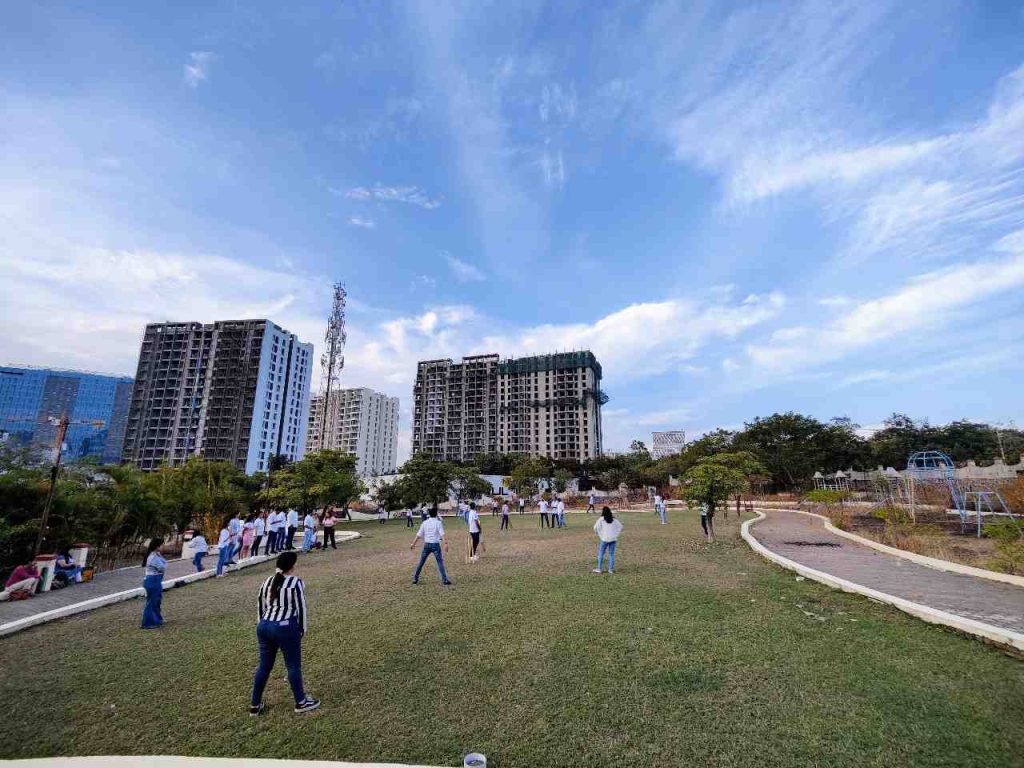 Large group of people playing outdoor games on open green lawn at Kapila Resorts Kharadi Pune with city skyline in background